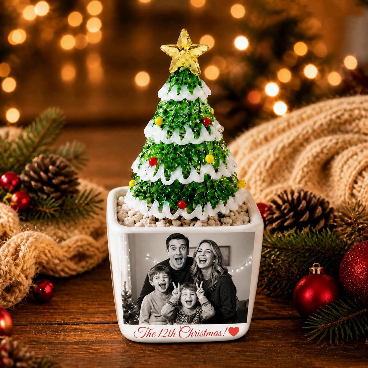 May include: A square white ceramic planter featuring a black and white family photograph and the text "The 12th Christmas!" with a red heart. A green glass Christmas tree with white snow-like accents and a yellow star sits on top of the planter.