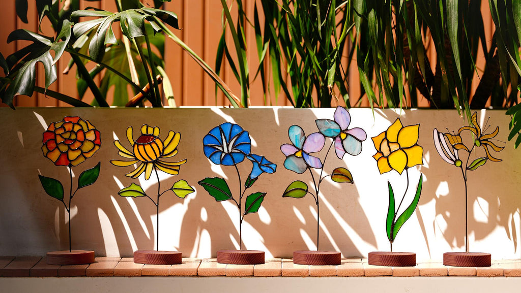 May include: A row of six colorful stained glass flower sculptures. Each flower has a unique design and color palette, including red, yellow, blue, and purple. The flowers are mounted on thin metal stems with brown circular bases.