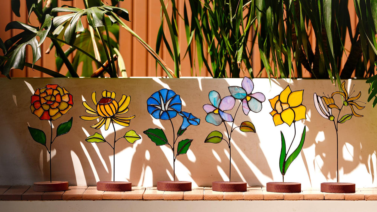 May include: A row of six colorful stained glass flower sculptures. Each flower has a unique design and color palette, including red, yellow, blue, and purple. The flowers are mounted on thin metal stems with brown circular bases.