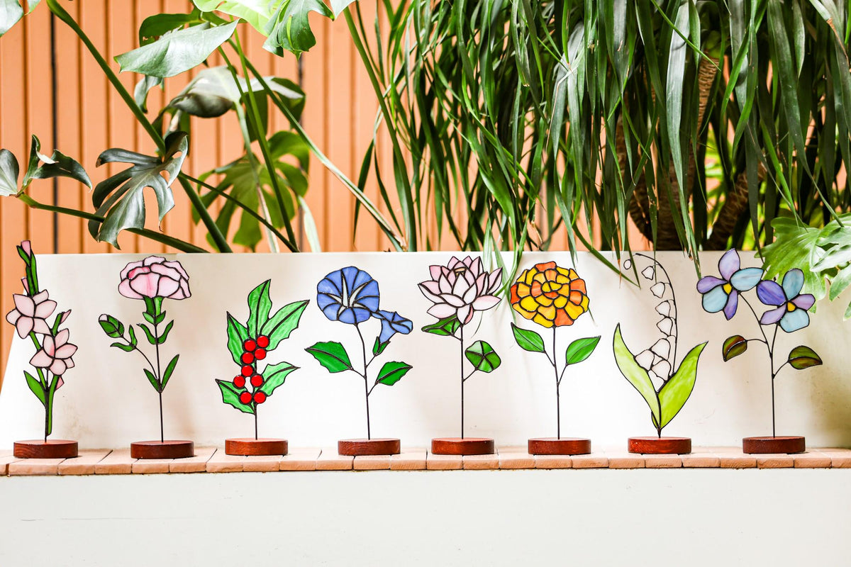 May include: A collection of colorful stained glass flower sculptures, each with a unique design and mounted on a small wooden base. The flowers include pink, blue, yellow, and white blooms.