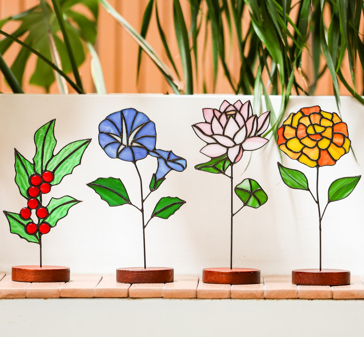 May include: Four stained glass flower sculptures. The first is a holly branch with red berries. The second is a blue morning glory. The third is a pink lotus flower. The fourth is a yellow and orange marigold. Each is mounted on a wooden base.