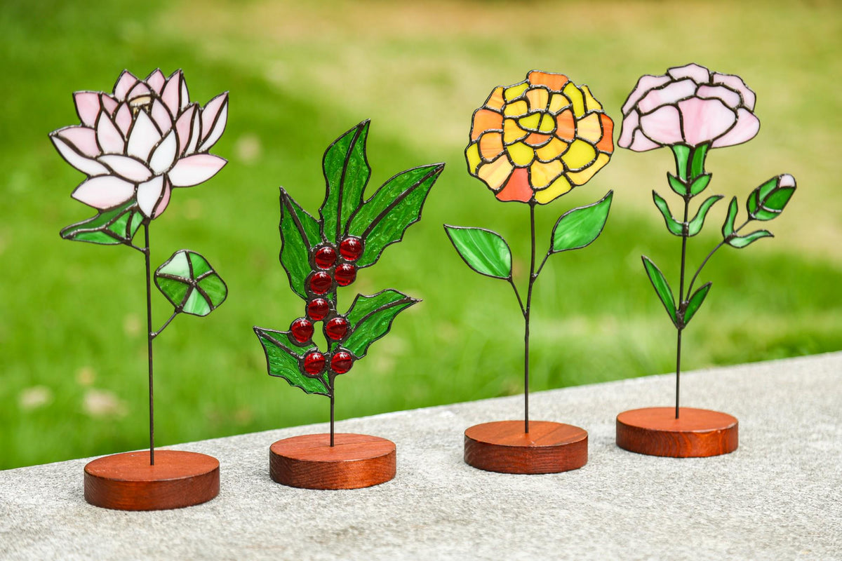May include: Four stained glass flower sculptures with wooden bases. The flowers include a pink and white lotus, a green holly branch with red berries, a yellow and orange flower, and a pink flower. The stems are thin and black.