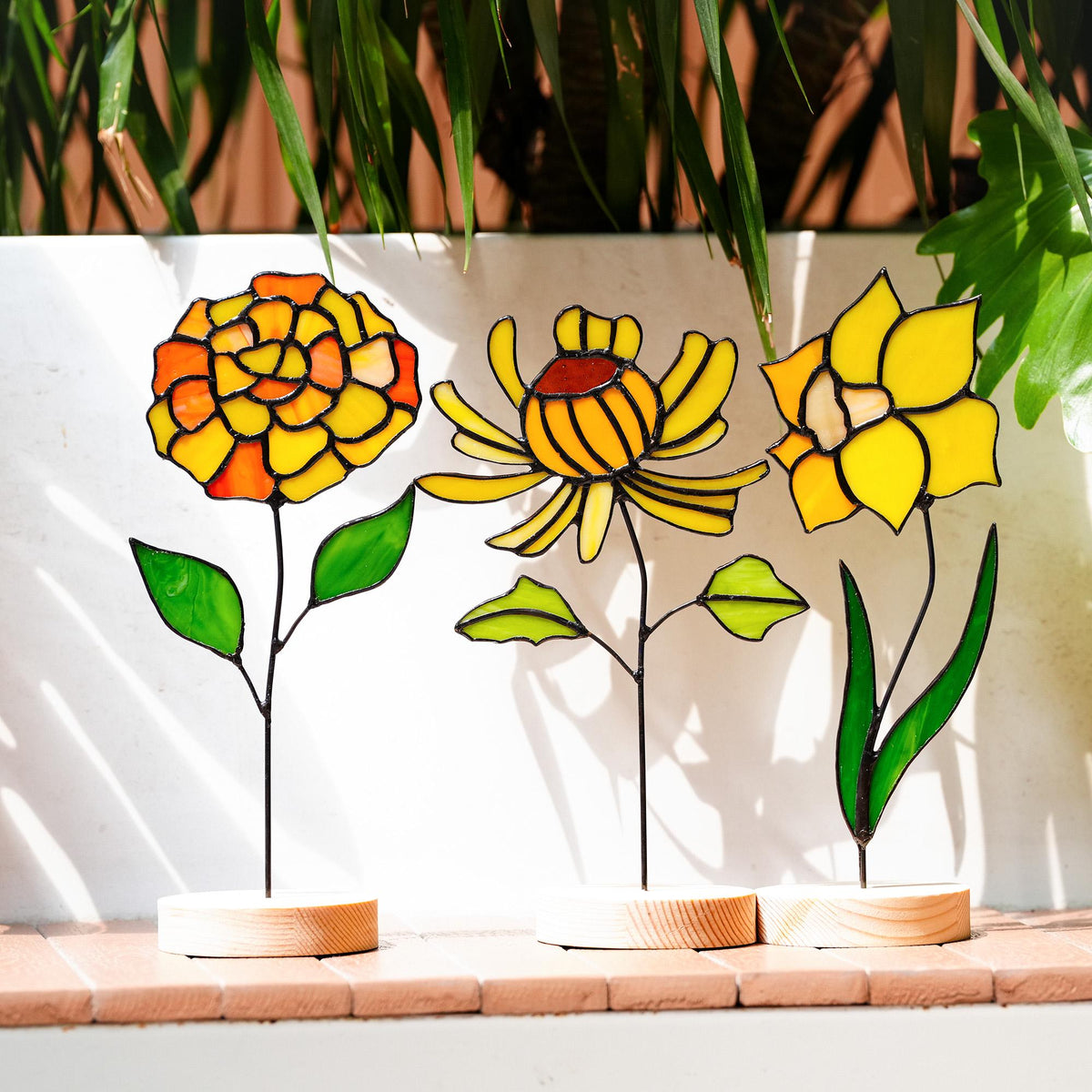 May include: Three stained glass flower sculptures. The flowers are yellow and orange with black outlines, and green leaves. Each flower is mounted on a thin black stem and set in a small, round wooden base. The background is a white wall and green plants.
