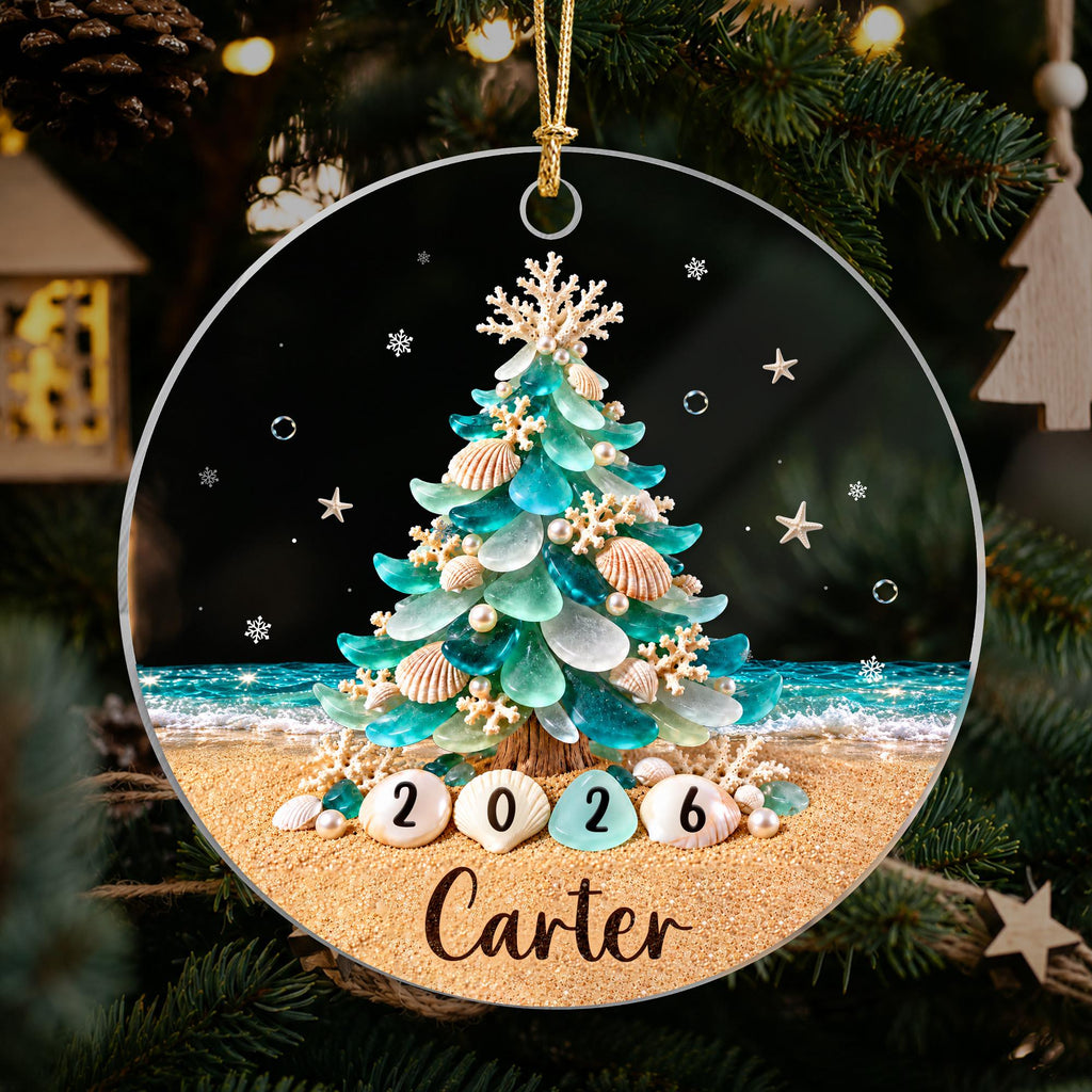 May include: A round Christmas ornament featuring a beach-themed tree made of sea glass, seashells, and coral. The tree sits on a sandy beach with ocean waves in the background. The year 2026 is printed on shells, and the name Carter is below.