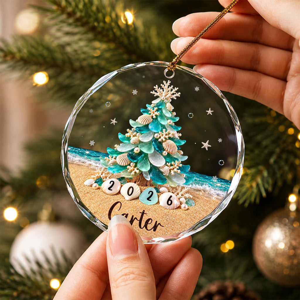 May include: A circular clear glass ornament featuring a printed design of a Christmas tree made from teal sea glass and white seashells on a sandy beach. The numbers 2026 are displayed on shells at the base, with the name Carter written below.