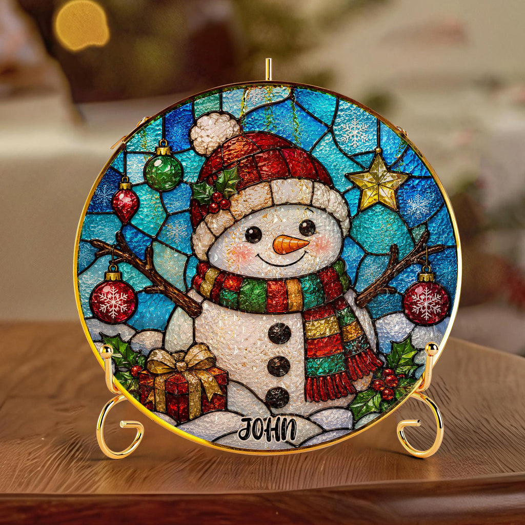 May include: A circular stained-glass style ornament featuring a snowman wearing a red hat and a multi-colored scarf. The background displays blue glass patterns with hanging ornaments, a star, and snowflakes. The name JOHN is printed at the bottom.