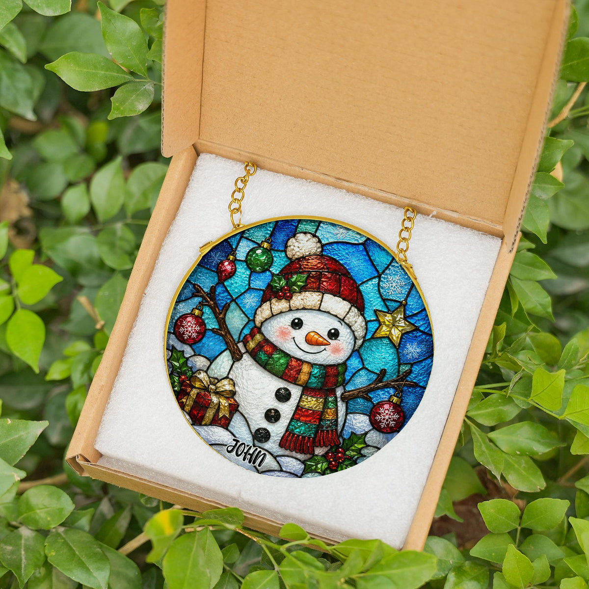 May include: A circular stained glass ornament featuring a snowman wearing a red hat and a multi-colored scarf, surrounded by blue geometric patterns, Christmas ornaments, and a gift box. The name JOHN is printed at the bottom of the ornament.