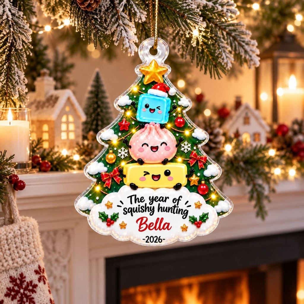 May include: A clear, tree-shaped Christmas ornament featuring a stack of three cartoon characters: a blue cube, a pink dumpling, and a yellow rectangle. The ornament includes the text "The year of squishy hunting Bella -2026-" and holly accents.