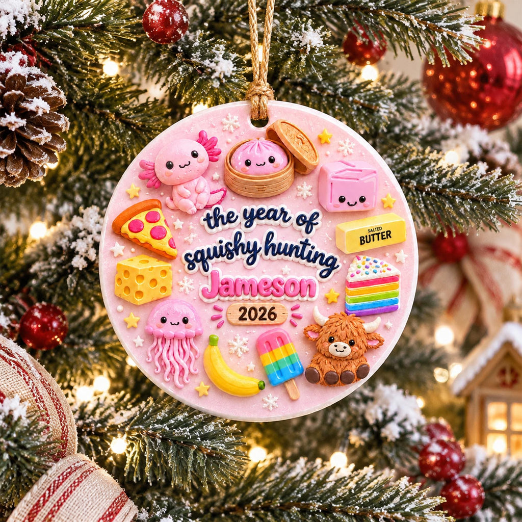 May include: A round, light pink Christmas ornament featuring a variety of colorful, cute, squishy-style figures including an axolotl, a dumpling, a slice of pizza, cheese, a jellyfish, a banana, a popsicle, and a highland cow. The text reads: the year of squishy hunting Jameson 2026.