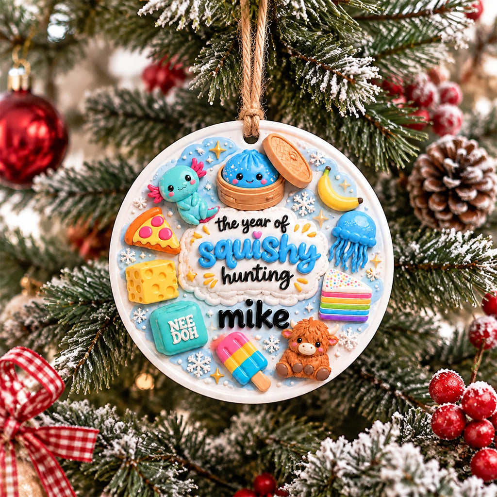 May include: A round white Christmas ornament hanging from a jute string, featuring raised colorful figures including an axolotl, a dumpling, a banana, a jellyfish, a slice of pizza, cheese, cake, a popsicle, and a highland cow with the text "the year of squishy hunting mike".
