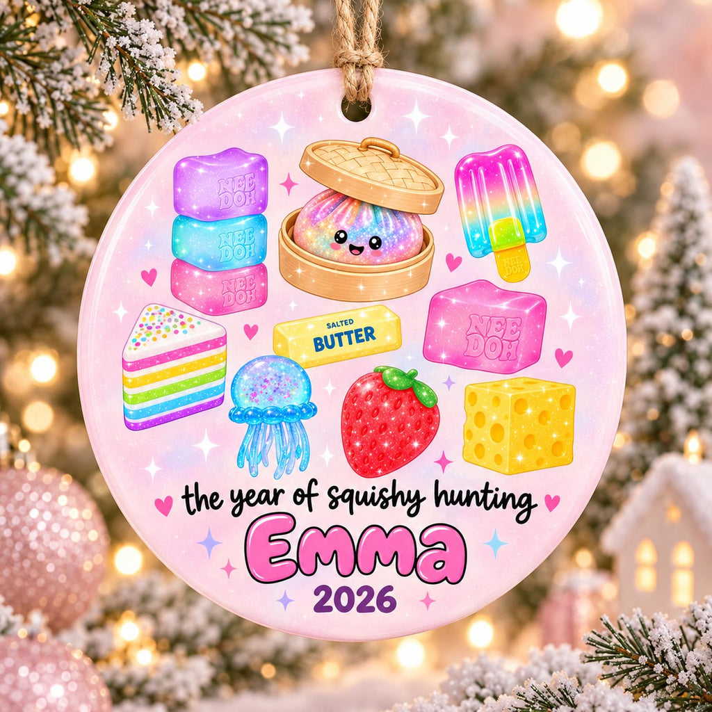 May include: A round ceramic ornament featuring a pastel pink background with illustrations of a rainbow cake, a jellyfish, a strawberry, a block of cheese, a popsicle, a dumpling in a steamer, and blocks labeled Nee Doh and Salted Butter. Text reads: the year of squishy hunting Emma 2026.