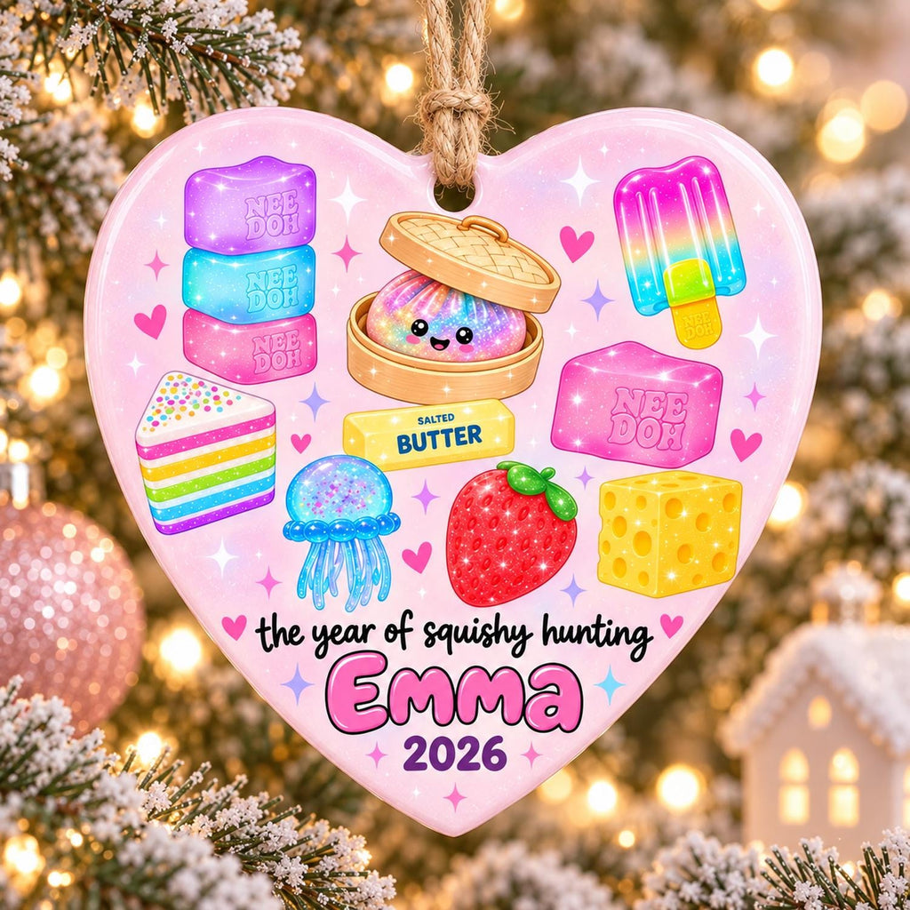 May include: A heart-shaped ornament with a light pink background featuring colorful illustrations of a rainbow cake, a jellyfish, a strawberry, cheese, a popsicle, and squishy toys. Text reads: the year of squishy hunting, Emma, 2026.