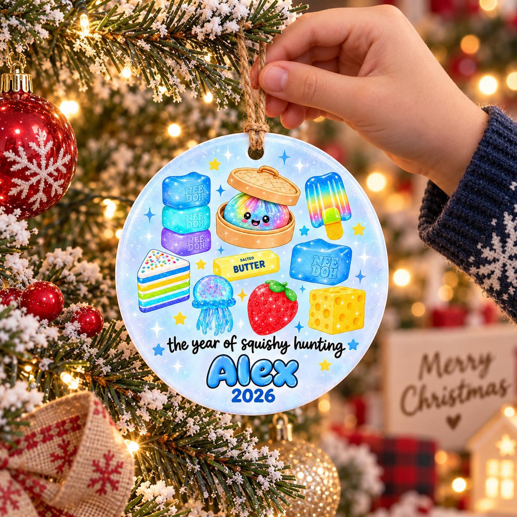 May include: A round, flat Christmas ornament featuring a colorful graphic of various squishy toys, including a dumpling, popsicle, cake, strawberry, and cheese. The text reads, "the year of squishy hunting," "Alex," and "2026" in blue and black lettering.
