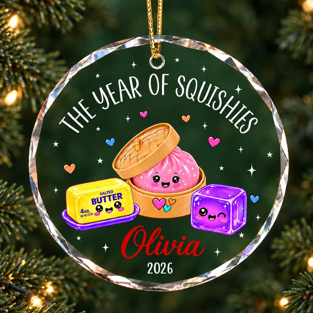 May include: A round, clear acrylic ornament featuring the text THE YEAR OF SQUISHIES, a cartoon pink dumpling in a bamboo steamer, a yellow stick of salted butter, and a purple jelly cube. The name Olivia and the year 2026 are printed below.