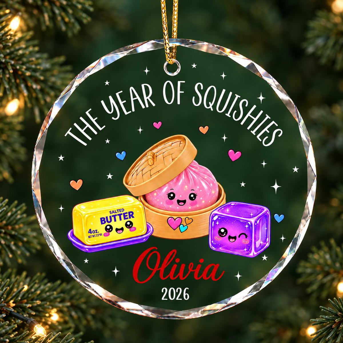 May include: A round, clear acrylic ornament featuring the text THE YEAR OF SQUISHIES, a cartoon pink dumpling in a bamboo steamer, a yellow stick of salted butter, and a purple jelly cube. The name Olivia and the year 2026 are printed below.