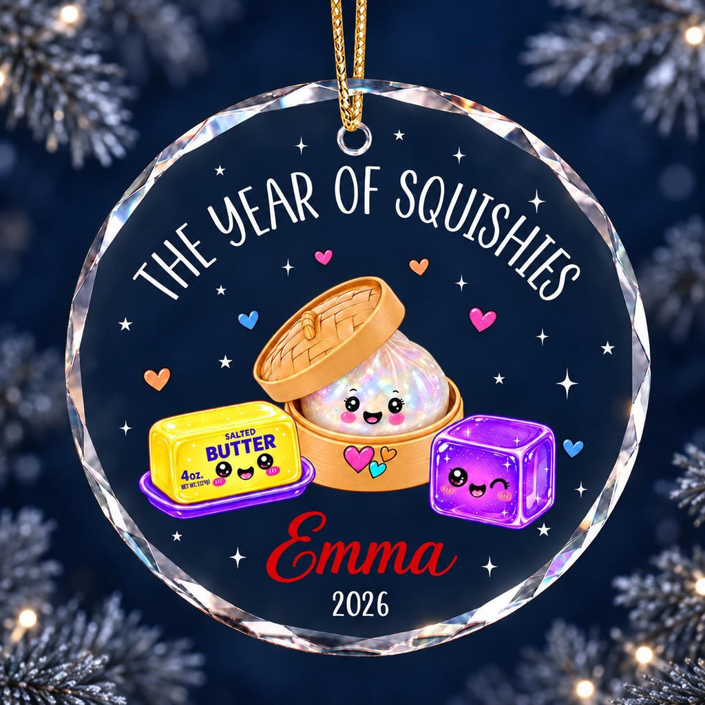 May include: A round, clear glass ornament featuring the text THE YEAR OF SQUISHIES, a cartoon dumpling in a bamboo steamer, a stick of salted butter labeled 4oz, and a purple cube. The name Emma and the year 2026 are printed in red and white.