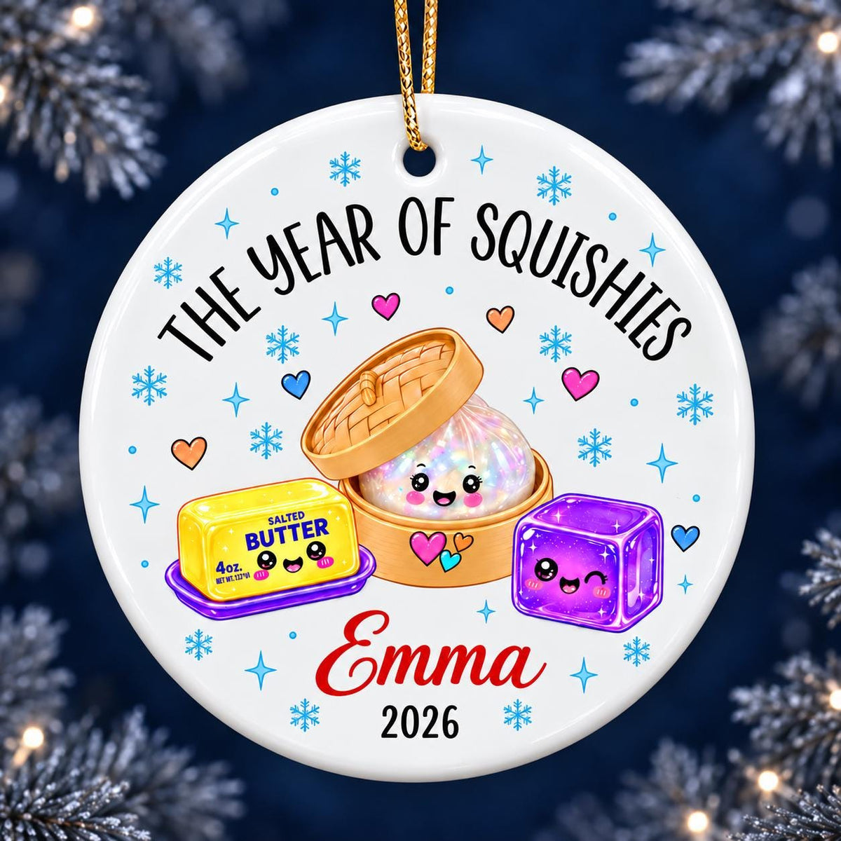 May include: A round white ceramic ornament featuring the text THE YEAR OF SQUISHIES, the name Emma, and the year 2026. The design includes a yellow stick of salted butter, a purple jelly cube, and a dumpling in a bamboo steamer, all with faces.