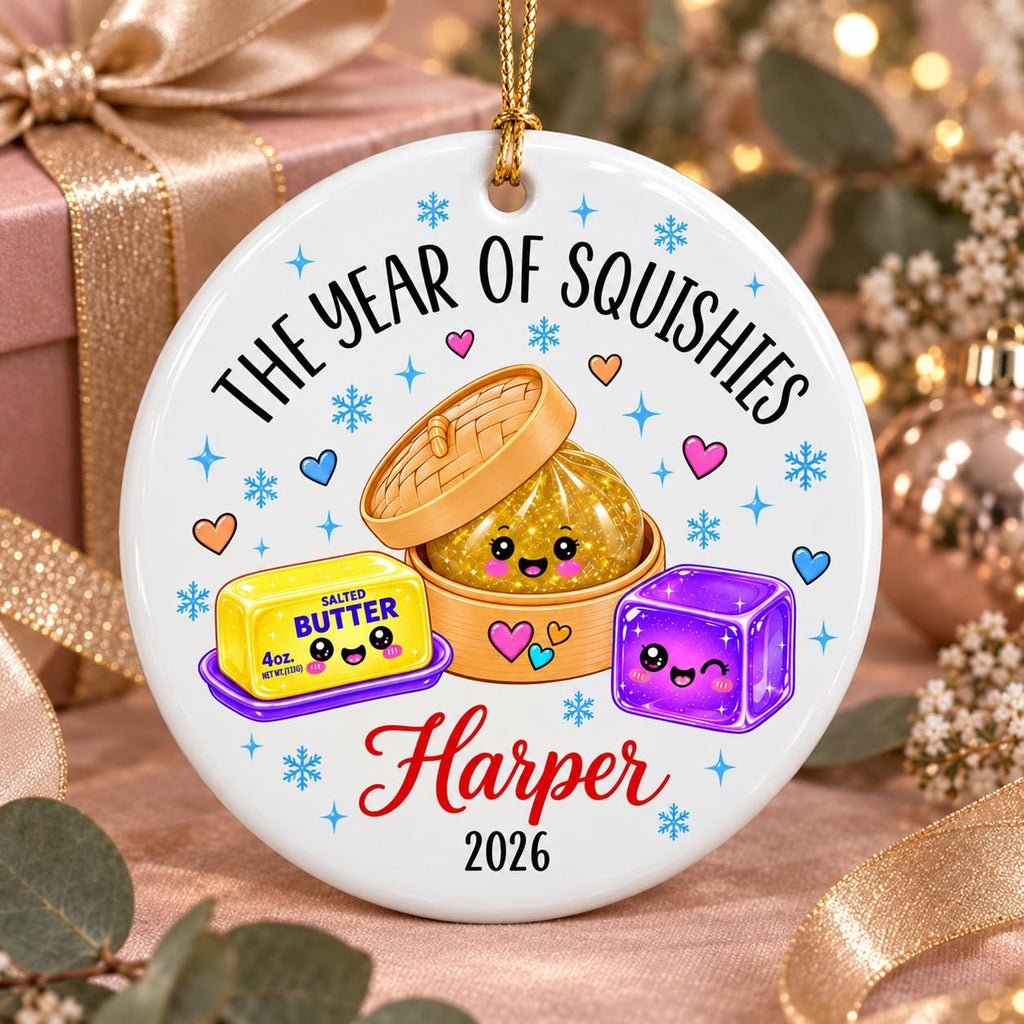 May include: A round white ceramic ornament featuring the text THE YEAR OF SQUISHIES, Harper, and 2026. The design includes a gold glitter dumpling in a bamboo steamer, a yellow stick of salted butter, and a purple cube, all with cute faces.