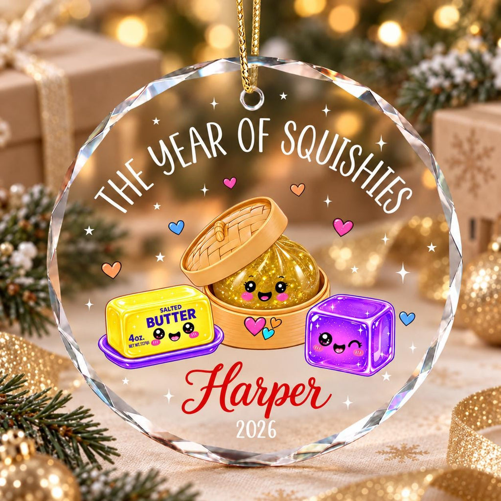 May include: A round, faceted glass ornament featuring the text THE YEAR OF SQUISHIES, the name Harper, and the year 2026. The design includes cartoon illustrations of a stick of butter, a dumpling in a steamer, and a purple cube with faces.