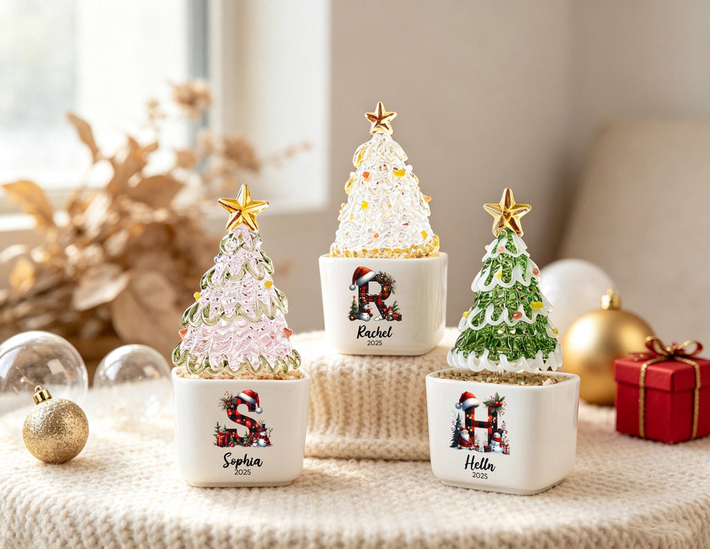 May include: Three decorative Christmas tree ornaments in white square ceramic pots. Each tree is made of textured glass topped with a gold star. The pots feature a plaid letter with a Santa hat, a name, and the year 2025 printed on the front.