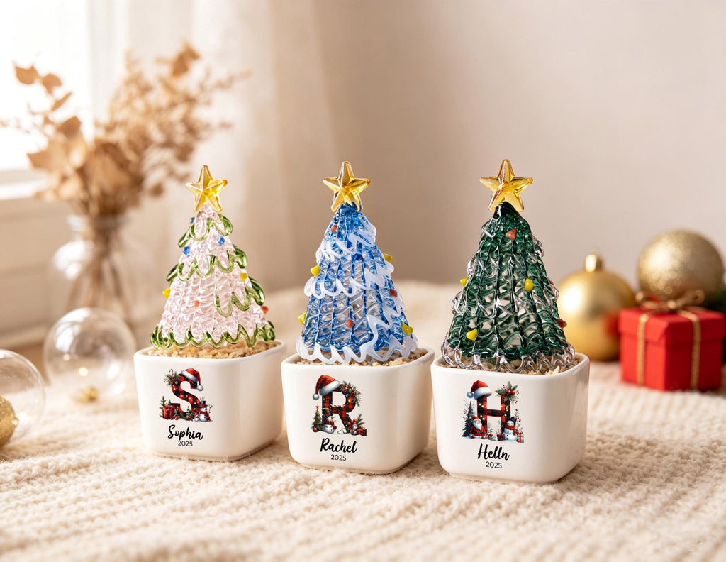 May include: Three glass Christmas tree ornaments in white square pots. The trees are pink, blue, and green with gold stars on top. Each pot features a letter S, R, or H decorated with a Santa hat, along with the names Sophia, Rachel, and Helln and 2025.