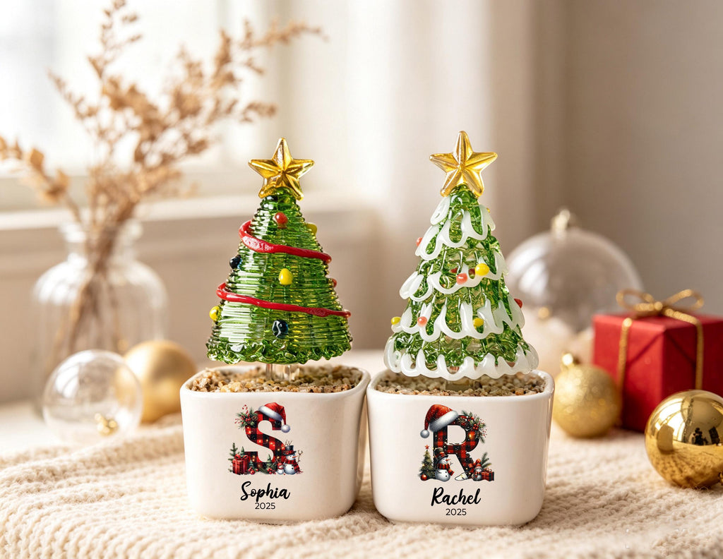 May include: Two glass Christmas tree ornaments in white square planters. The left tree has red garland and the letter S with the name Sophia and 2025. The right tree has white garland and the letter R with the name Rachel and 2025. Both have gold stars.