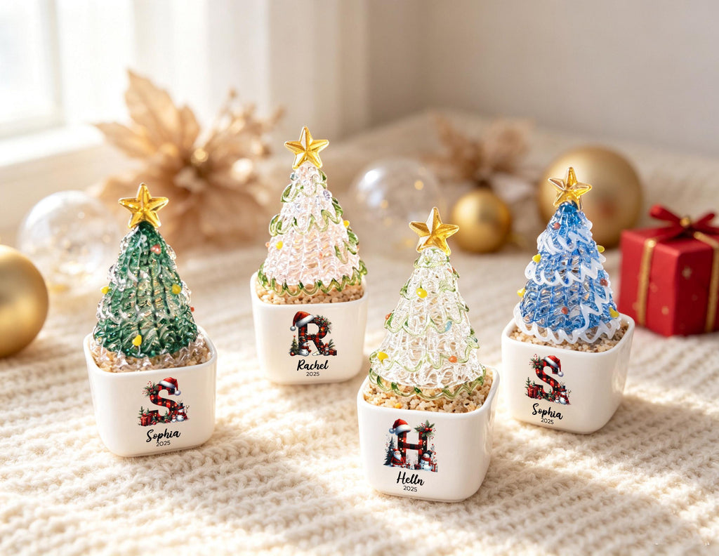 May include: Four small, conical, textured glass Christmas trees in white square ceramic pots. Each tree is topped with a gold star and features a personalized letter with a Santa hat, a name, and the year 2025 printed on the front of the pot.