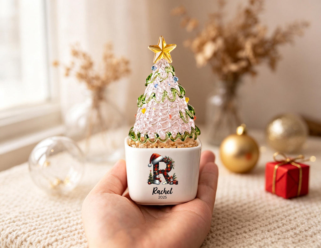 May include: A small glass Christmas tree ornament with a gold star topper, sitting in a white square ceramic planter. The planter features a graphic of the letter R in a red plaid pattern with a Santa hat, the name Rachel, and the year 2025.