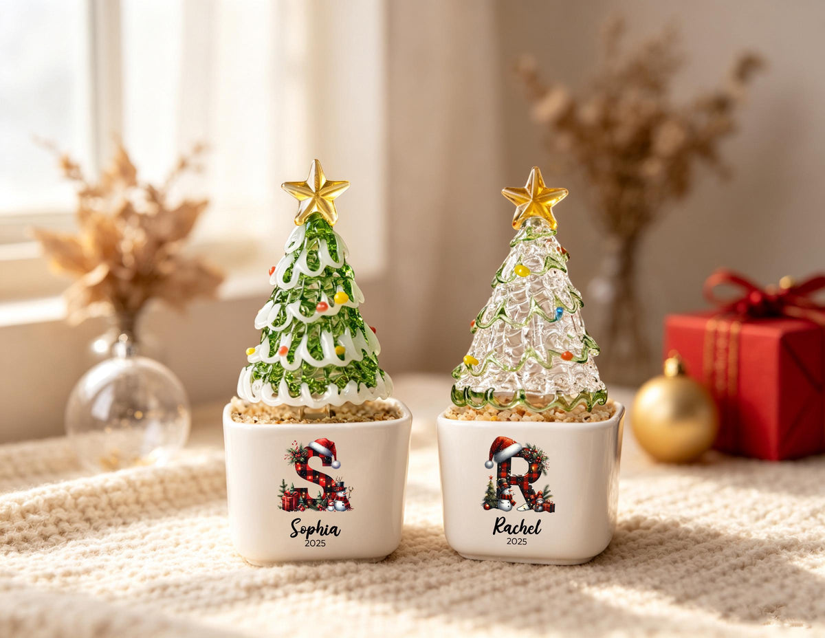 May include: Two glass Christmas tree ornaments in white square planters. The left tree is green with a gold star, featuring the letter S, the name Sophia, and 2025. The right tree is clear with a gold star, featuring the letter R, the name Rachel, and 2025.