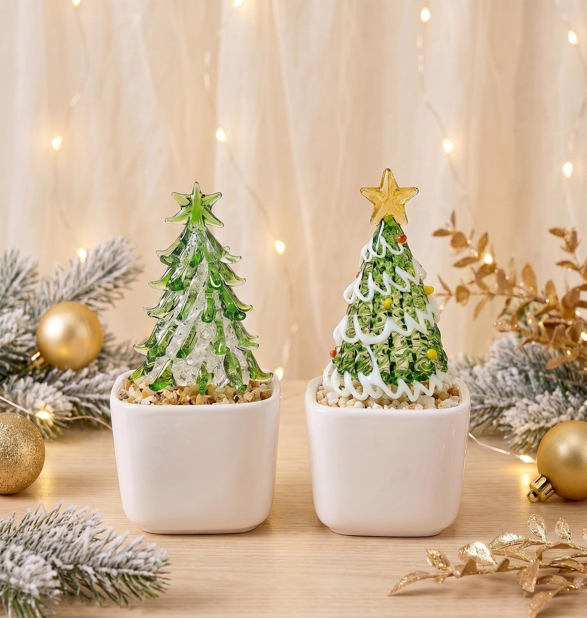 May include: Two glass Christmas tree figurines in white square ceramic pots. The left tree is green with a green star topper. The right tree is green with white swirling accents, colorful dots, and a yellow star topper. Both sit on small pebbles.
