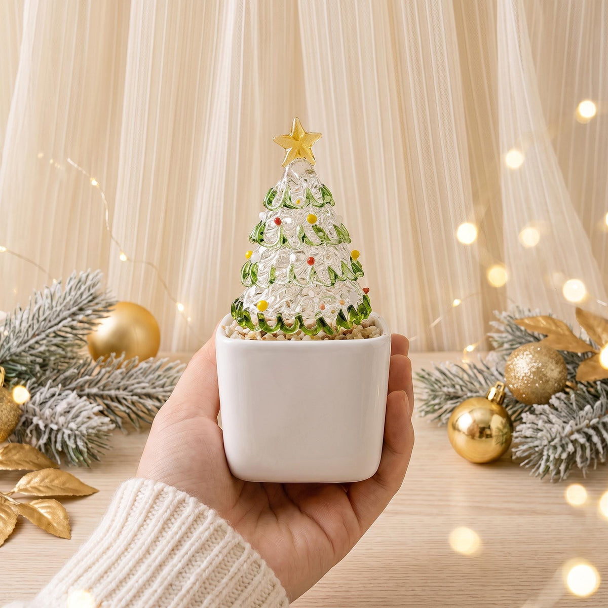 May include: A clear glass Christmas tree figurine with green glass accents and small colorful glass dots, topped with a gold star. The tree is set in a square white ceramic planter filled with small light-colored pebbles.