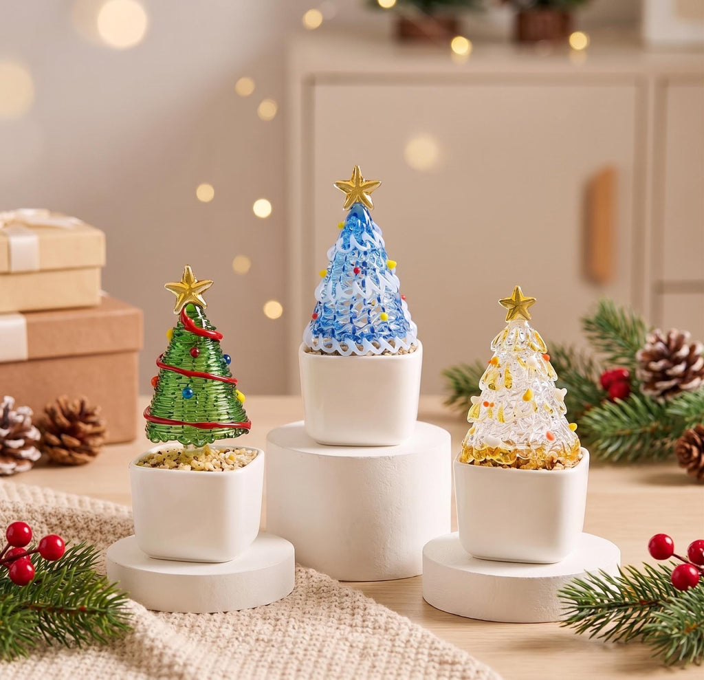May include: Three decorative glass Christmas trees in white square pots. One tree is green with red garland, one is blue with white swirls, and one is clear with gold accents. Each tree is topped with a small gold star and sits on a white base.