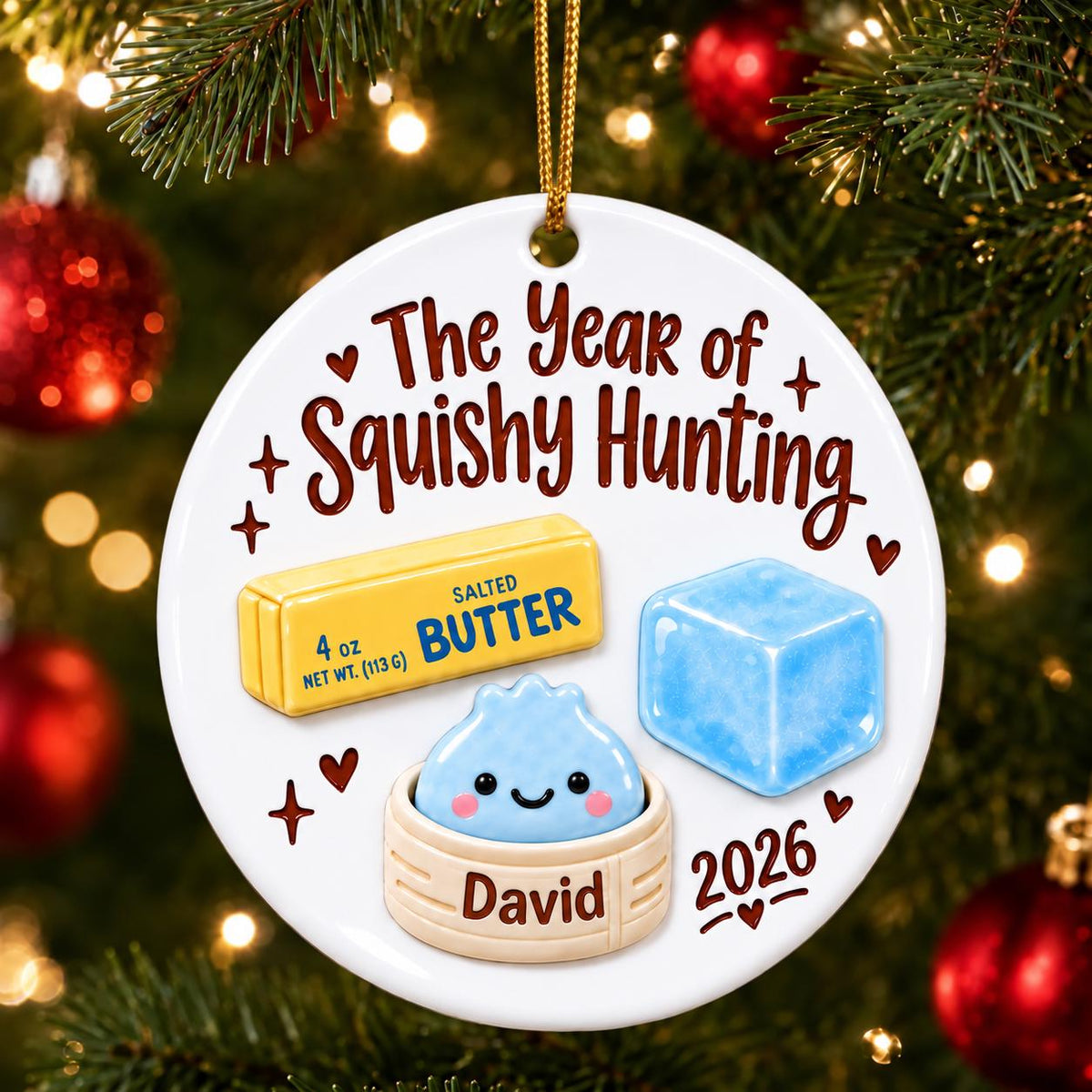 May include: A round white ceramic ornament featuring the text The Year of Squishy Hunting and 2026. The design includes a yellow stick of salted butter labeled 4 oz, a blue ice cube, and a light blue dumpling in a steamer basket labeled David.