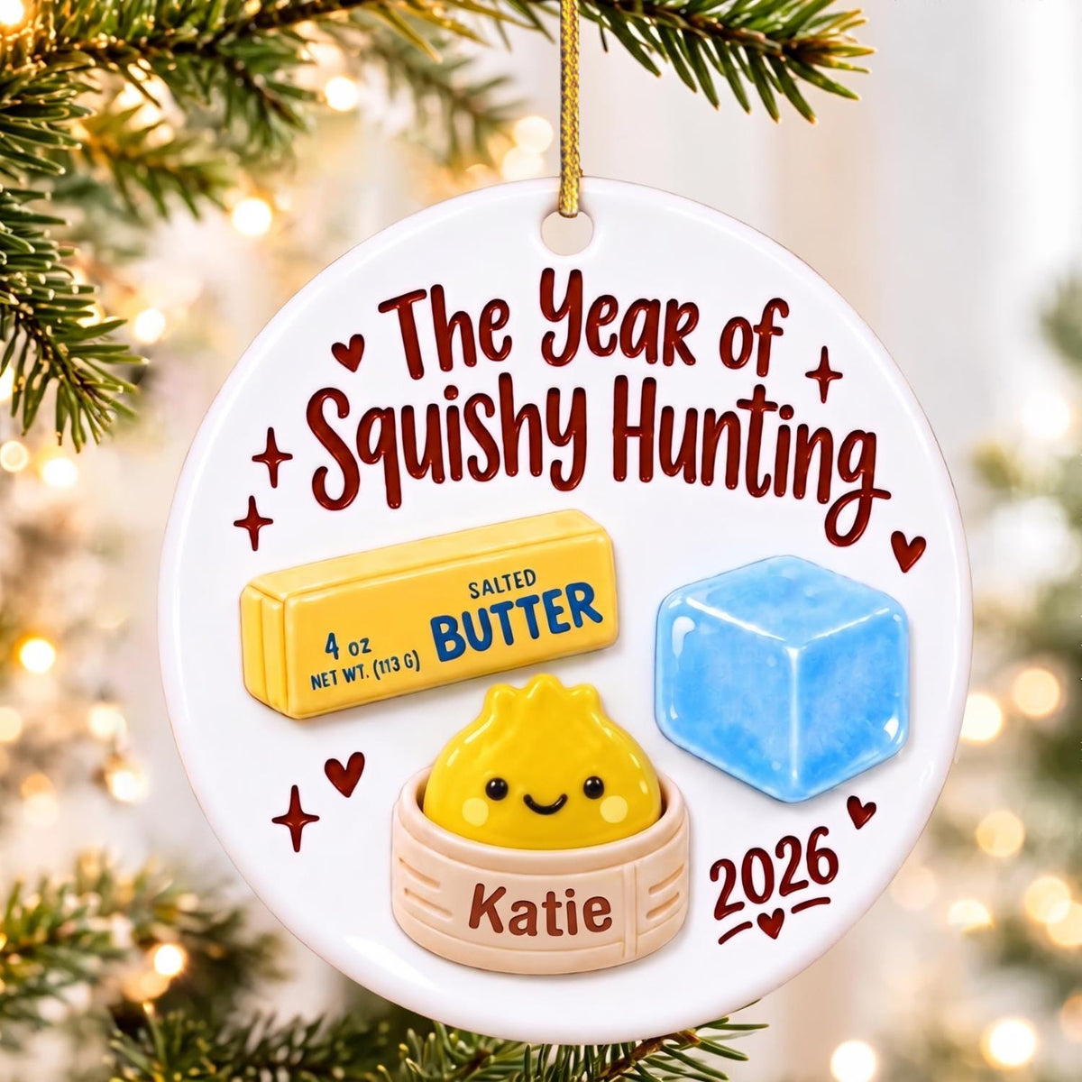 May include: A round white ceramic ornament with a gold string featuring the text The Year of Squishy Hunting and 2026. It displays a yellow stick of salted butter labeled 4 oz, a blue ice cube, and a yellow dumpling in a steamer basket labeled Katie.
