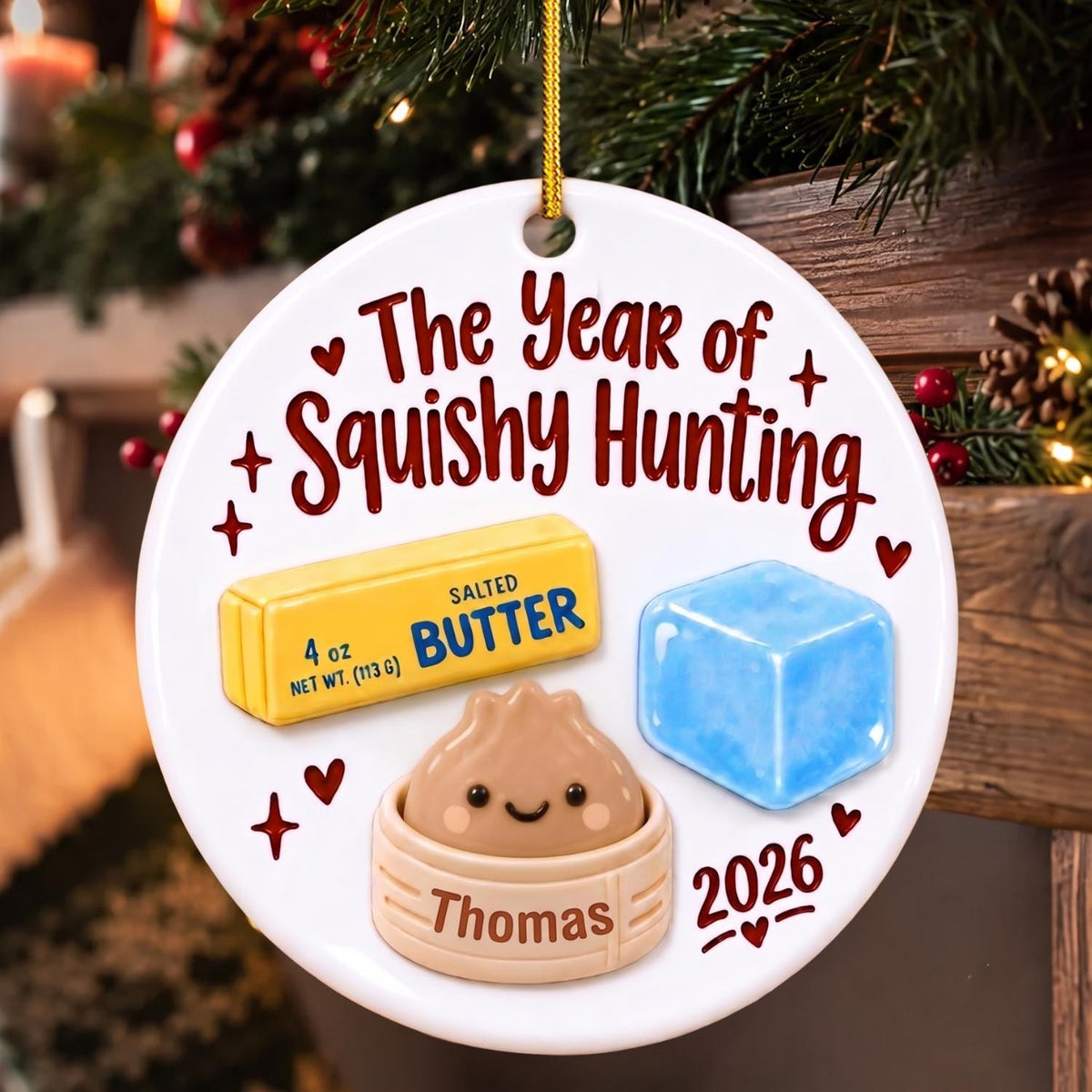 May include: A round white ceramic ornament featuring the text The Year of Squishy Hunting, a yellow stick of salted butter, a blue ice cube, and a tan dumpling in a steamer basket labeled Thomas, with the year 2026 printed in red at the bottom.