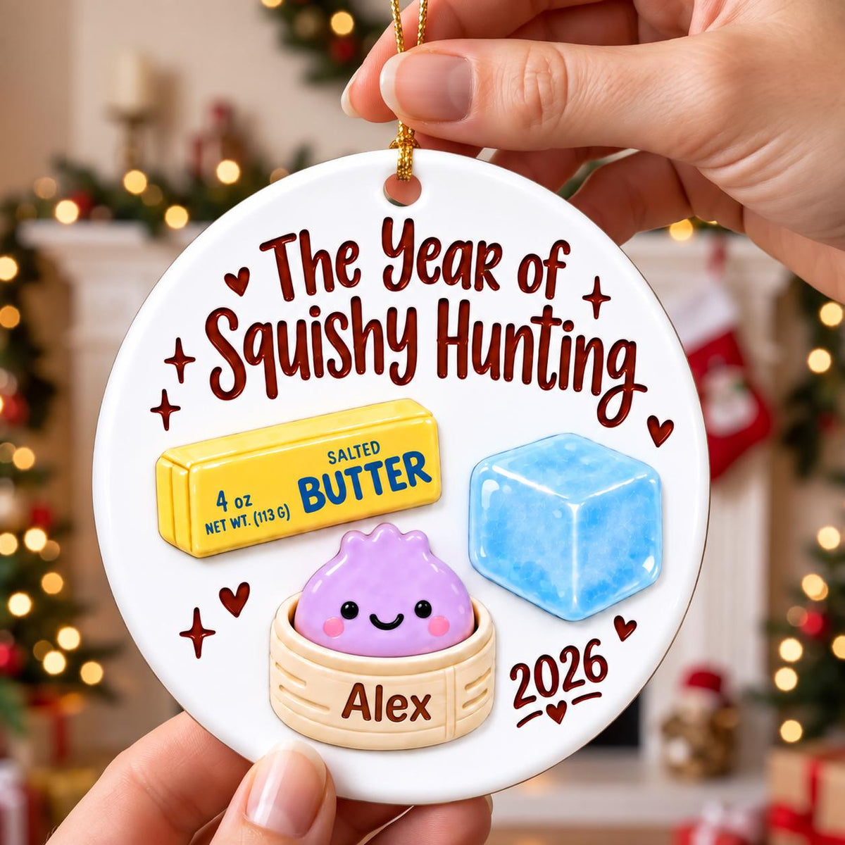 May include: A round white ceramic ornament featuring the text The Year of Squishy Hunting, a yellow stick of butter labeled 4 oz, a blue ice cube, and a purple dumpling in a bamboo steamer labeled Alex, with the year 2026 and small red hearts.