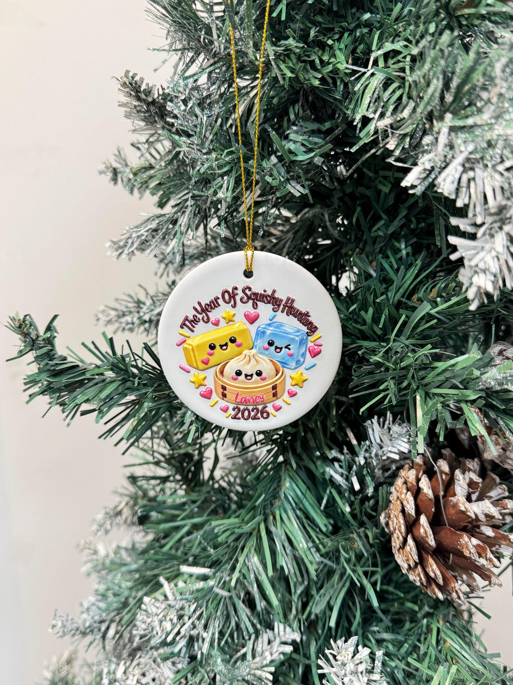 May include: A round white ceramic ornament hanging by a gold string. The design features a yellow butter stick, a blue ice cube, and a dumpling in a bamboo steamer, all with faces. Text reads The Year Of Squishy Hunting, Laney, and 2026.