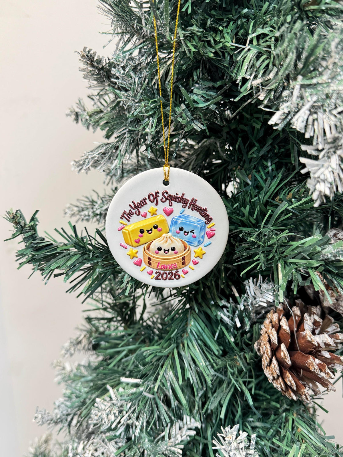 May include: A round white ceramic ornament hanging by a gold string. The design features a yellow butter stick, a blue ice cube, and a dumpling in a bamboo steamer, all with faces. Text reads The Year Of Squishy Hunting, Laney, and 2026.