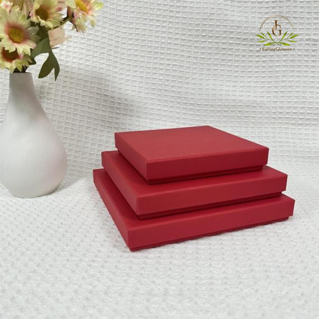 May include: A stack of three square red gift boxes with lids, arranged in descending order of size. The boxes have a smooth, matte finish and are positioned next to a white ceramic vase containing light pink and yellow flowers.