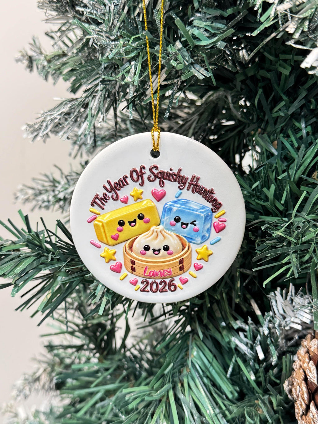 May include: A round white ceramic ornament hanging by a gold string, featuring a colorful graphic of a yellow block, a blue cube, and a dumpling in a bamboo steamer, all with smiling faces. The text reads The Year Of Squishy Hunting, Lames, 2026.