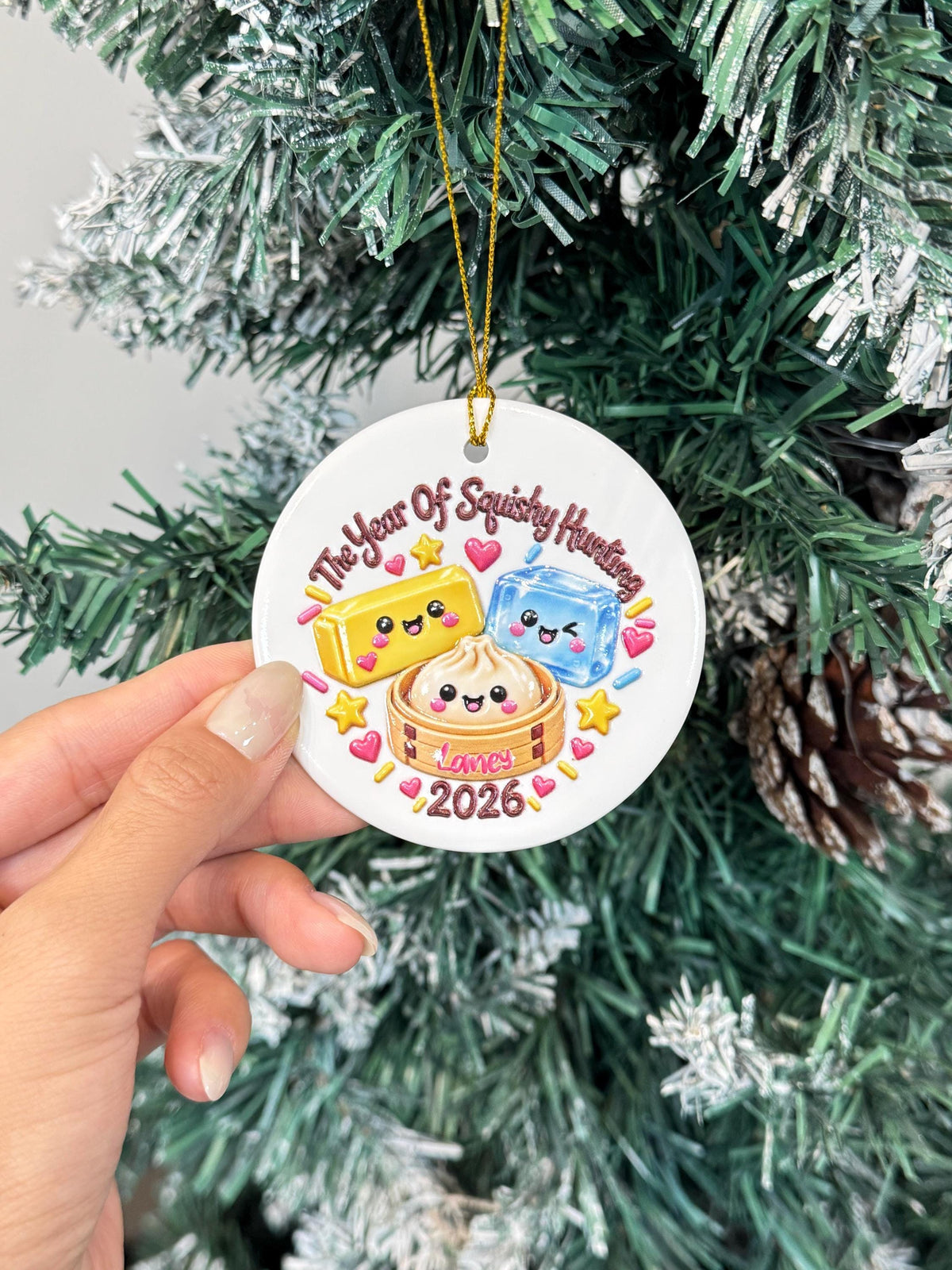 May include: A round white ceramic ornament featuring a cartoon illustration of a yellow block, a blue cube, and a dumpling in a bamboo steamer. The text reads The Year Of Squishy Hunting, Lamey, and 2026, surrounded by colorful hearts and stars.