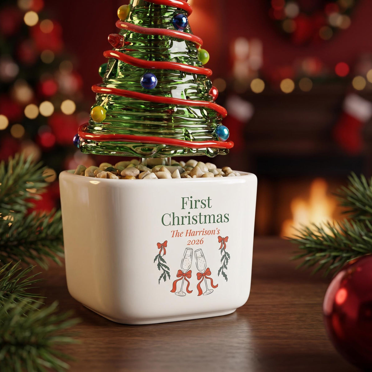 May include: A green glass Christmas tree ornament with red garland and colorful spheres, set in a white square ceramic planter filled with small stones. The planter features the text First Christmas, The Harrison's, 2026, and two champagne flutes.