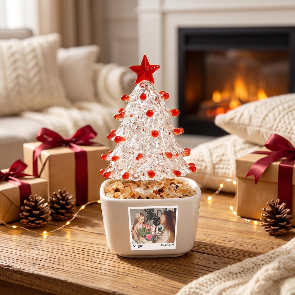 May include: A clear glass Christmas tree ornament with red accents and a red star topper, set in a white square planter filled with small stones. The planter features a square photo print of a person with the text Millie and 09.12.2036.