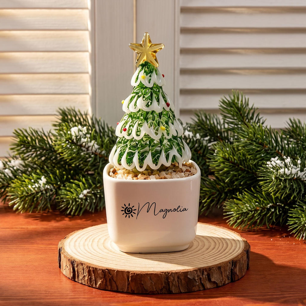 May include: A glass Christmas tree ornament with green and white swirled branches, topped with a gold star, and decorated with small colorful dots. The tree sits in a white square ceramic pot labeled with the word Magnolia and a sun icon.