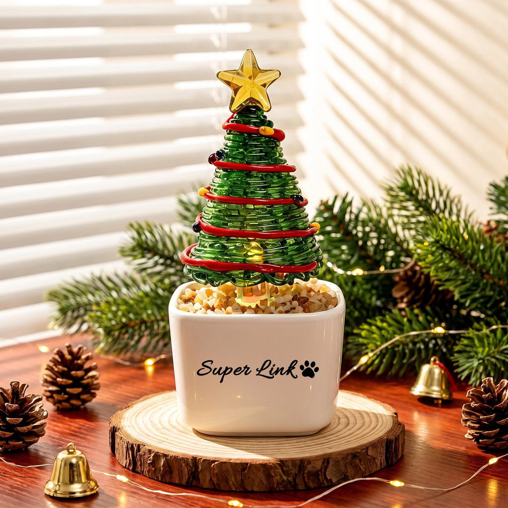 May include: A green glass spiral Christmas tree ornament with red glass garland and a yellow star topper, set in a white square ceramic pot labeled Super Link with a paw print. The pot contains small light-colored stones at the base.