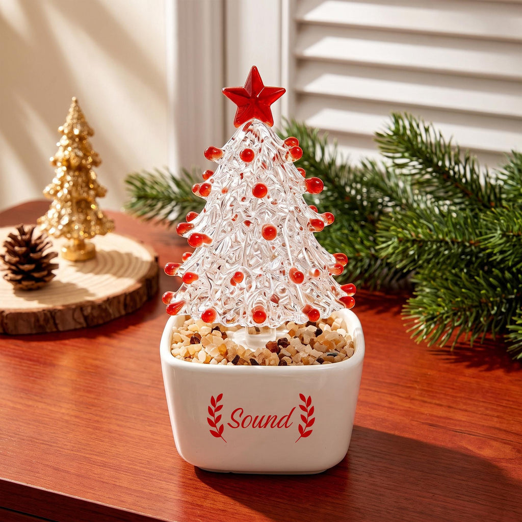 May include: A clear glass Christmas tree ornament with red glass accents and a red star topper, set in a white square ceramic planter filled with small tan stones. The planter features the red text "Sound" flanked by two red leaf sprigs.