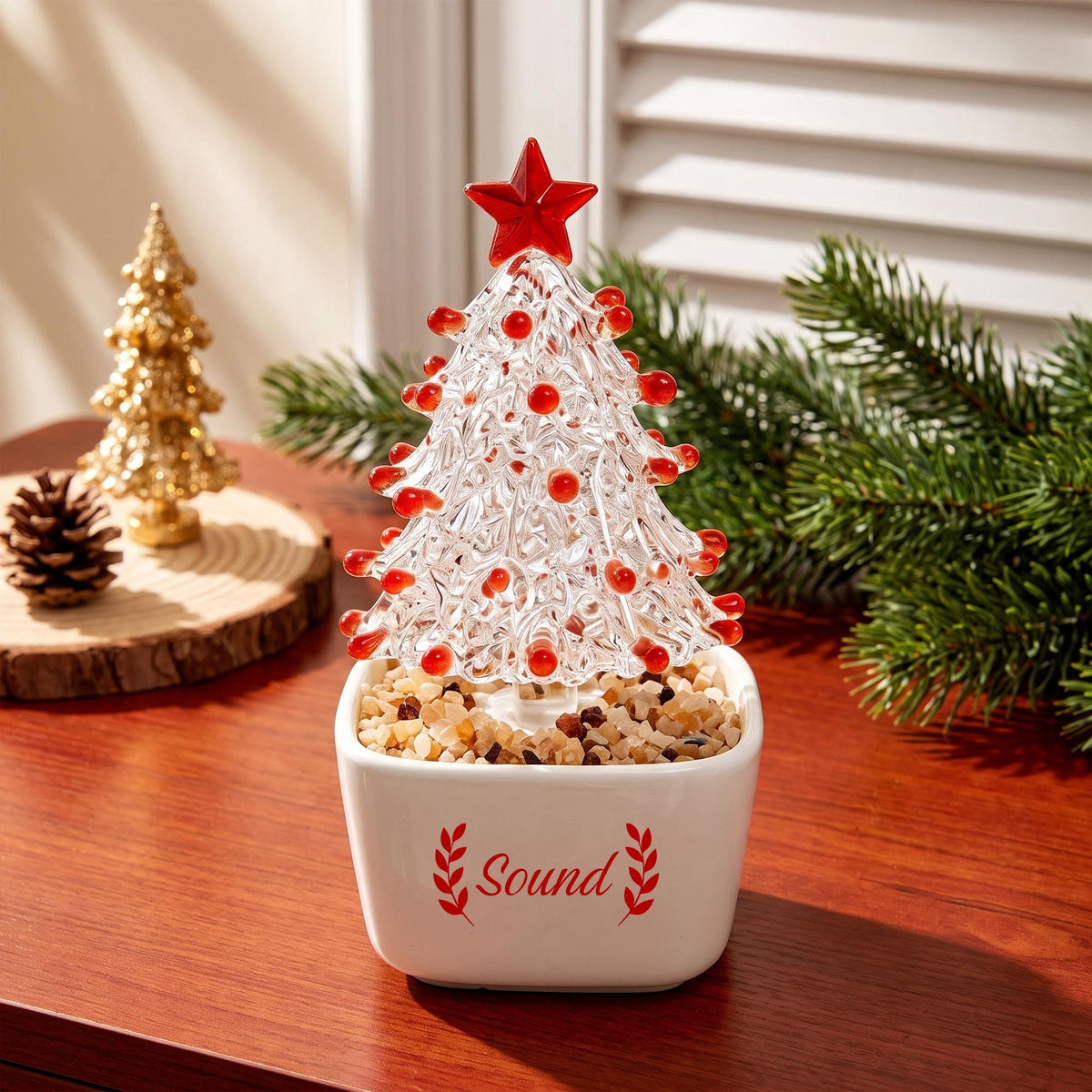 May include: A clear glass Christmas tree ornament with red glass accents and a red star topper, set in a white square ceramic planter filled with small tan stones. The planter features the red text "Sound" flanked by two red leaf sprigs.