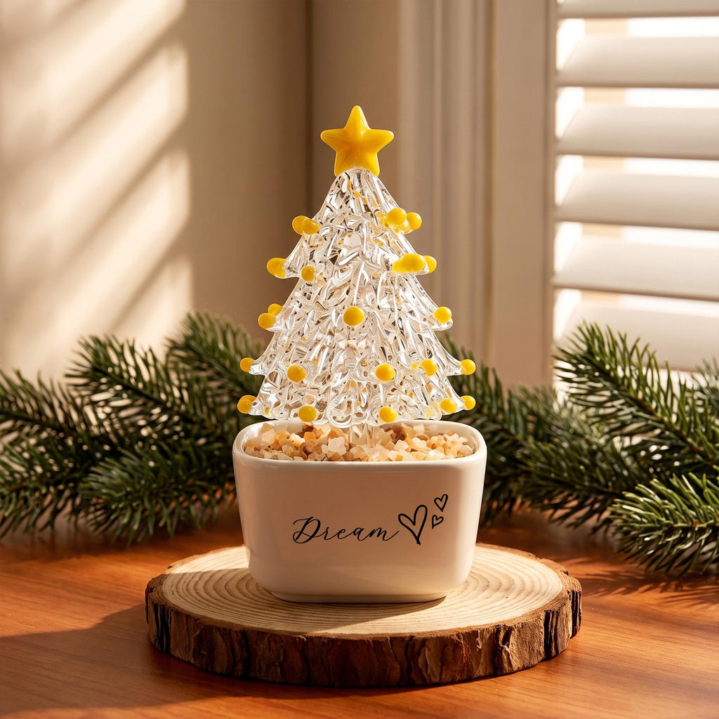May include: A clear glass Christmas tree figurine with yellow glass accents and a yellow star topper, set in a white ceramic pot filled with small stones. The pot features the black text "Dream" followed by a heart and two small dots.