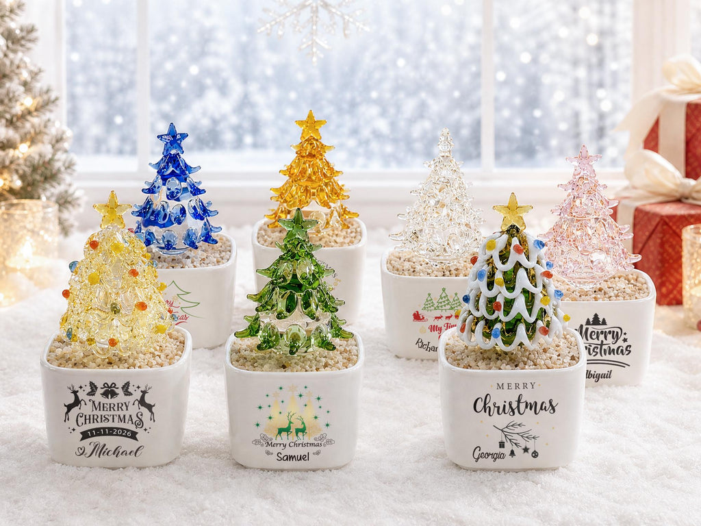 May include: A collection of seven miniature glass Christmas trees in various colors, including blue, yellow, green, clear, and pink. Each tree is topped with a star and sits in a white square ceramic planter filled with small pebbles and text.