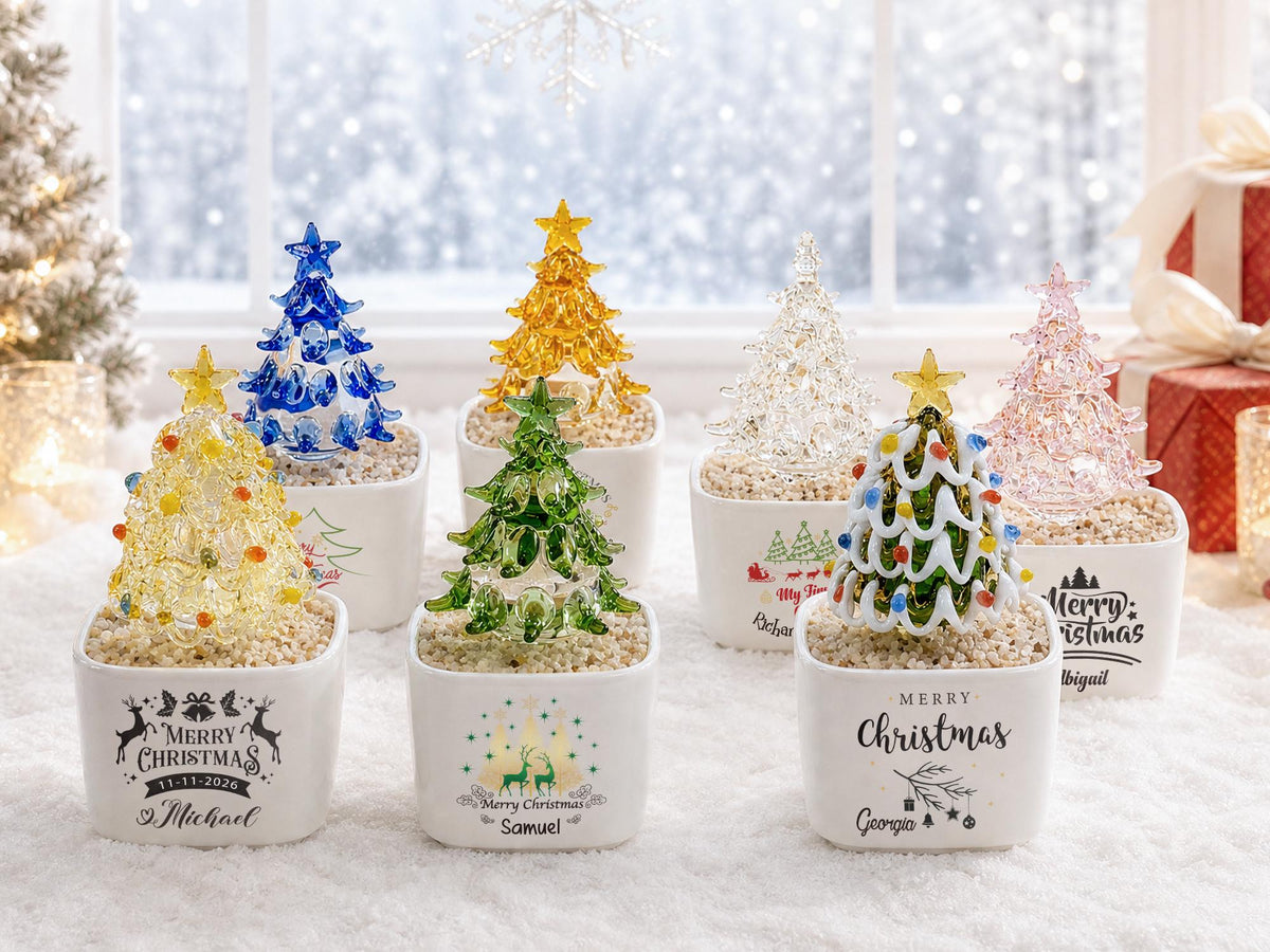 May include: A collection of seven miniature glass Christmas trees in various colors, including blue, yellow, green, clear, and pink. Each tree is topped with a star and sits in a white square ceramic planter filled with small pebbles and text.